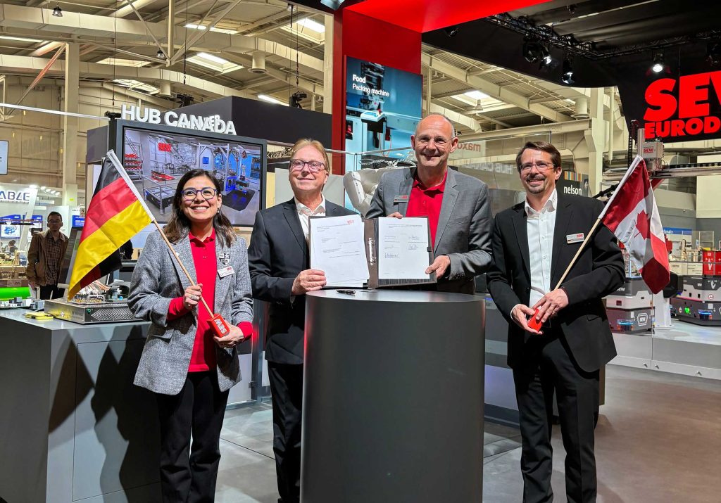 Four professionals at a trade show booth pose with documents and flags (Germany and Canada) in front of a 'HUB CANADA' display.