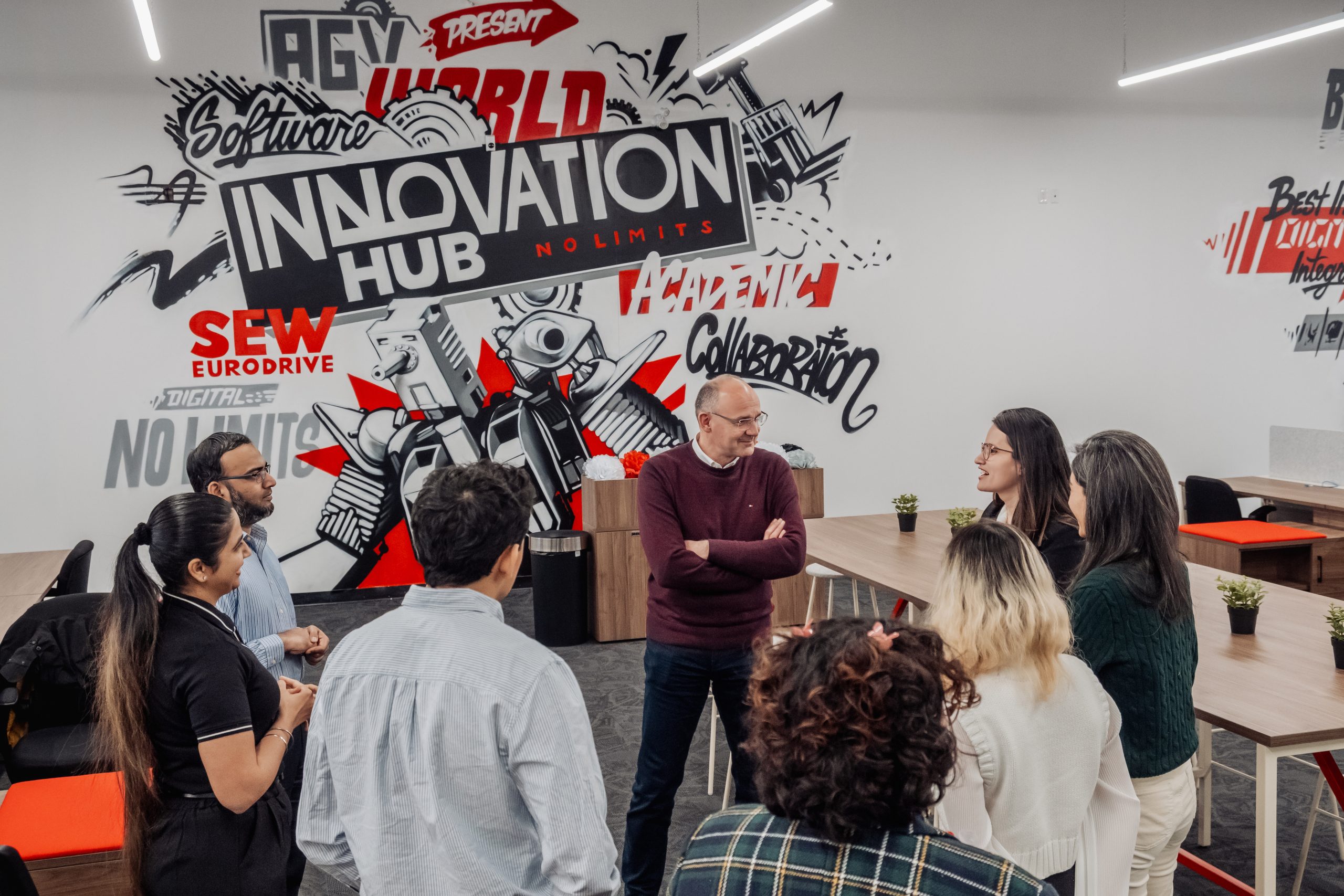 A group of colleagues stands in a modern coworking space, discussing around a wooden table near a colorful wall mural that reads 'INNOVATION HUB'.